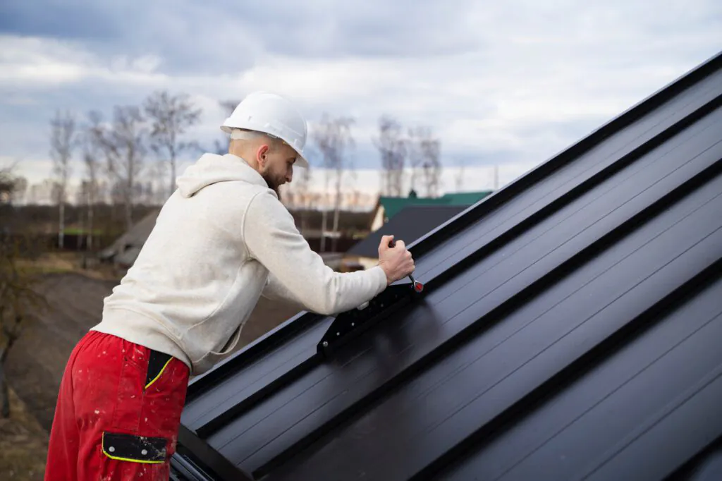 Roofer installing black metal roofing while wearing bright red pants